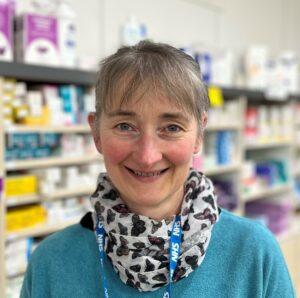 Pharmacist Julie Grigg is looking forward at the camera. She is wearing a blue top and a printed scarf. Behind her are the shelves of the pharmacy.
