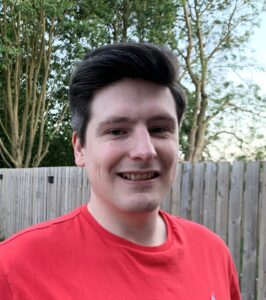 Dr Oliver Blow smiles at the camera. He is wearing a red t-shirt and there are trees behind him.
