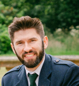 Dr Perry Sissons looks at the camera. He has short brown hair and a beard and is wearing a green tie and a dark blue jacket.