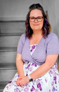 Dr Natalie Jones. She has mid length dark curly hair and glasses. She is standing in a GP practice and is wearing a green dress and jacket.