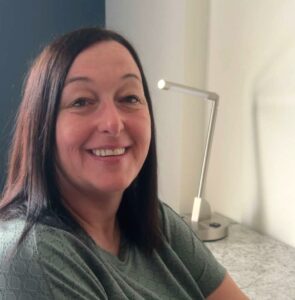 Claire Banks Practice Manager, Broughton House Surgery, looks at the camera. She has mid-length dark hair and is wearing a green shirt.