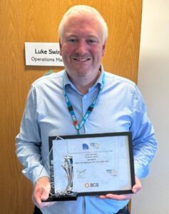 Luke Swindon, Operations Manager at Ossett Surgery holds an award certificate up to the camera. He wears a blue shirt.