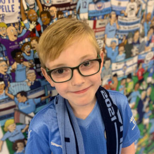 Mike, a 10 year old boy, looks at the camera wearing his favourite Manchester City football top with a background of Manchester City memorabilia