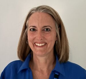 Caroline is smiling directly at the camera. She has shoulder length dark blonde hair and is wearing a royal blue shirt and NHS lanyard around her neck. She is standing in front of a plain light background