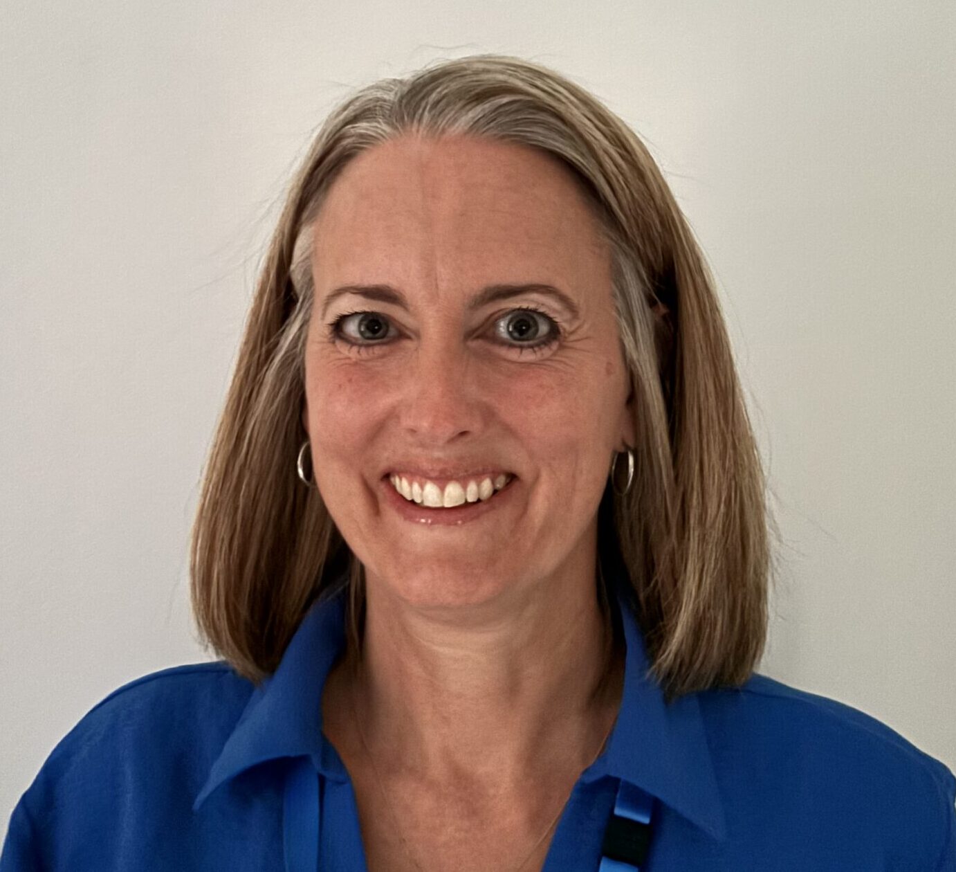 Caroline is smiling directly at the camera. She has shoulder length dark blonde hair and is wearing a royal blue shirt and NHS lanyard around her neck. She is standing in front of a plain light background