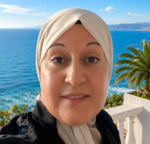 Naz is wearing a beige hijab and black top taking a selfie on a balcony overlooking a blue sea with distant mountains under a clear sky. There are green plants with pink flowers and a palm tree behind her.