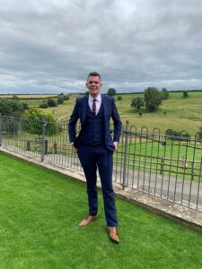 Photograph shows a full length image of a man in a blue suit standing outdoors in a green landscape with hills. The man is raising awareness about knowing the signs of testicular cancer and the importance of checking for lumps. 