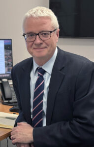 Picture of a man (Dr Trevor-Smith) is seated at a desk in an office. 