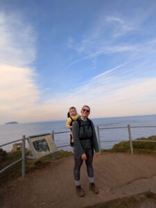 A woman with her baby daughter in a carrier on her back while she goes for a walk by the sea. 