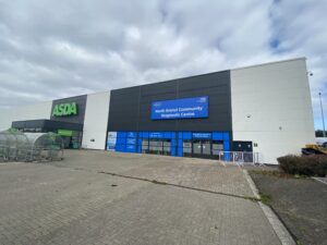 Photograph shows a building with a blue sign above the door with the words North Bristol Community Diagnostic Centre and the NHS logo in the top right hand corner. The doors are also blue and the building next door is an Asda supermarket.