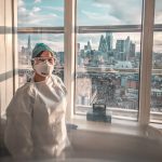 Drawn illustration of a NHS worker, in full personal protective equipment, stood in front of window