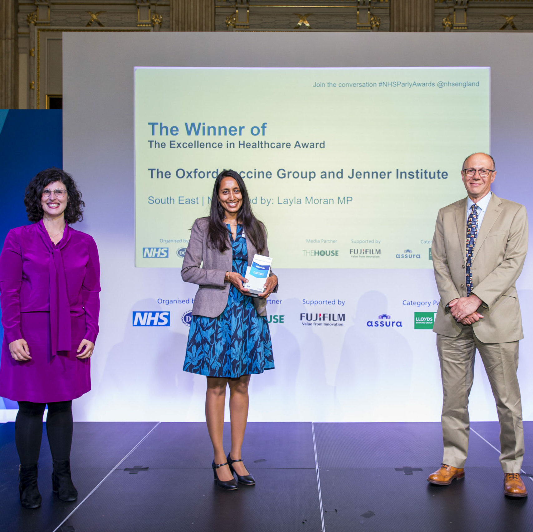 Three people are stood on the stage with a member of The Oxford Vaccine Group and the Jenner Institute receiving their Parliamentary NHS Award