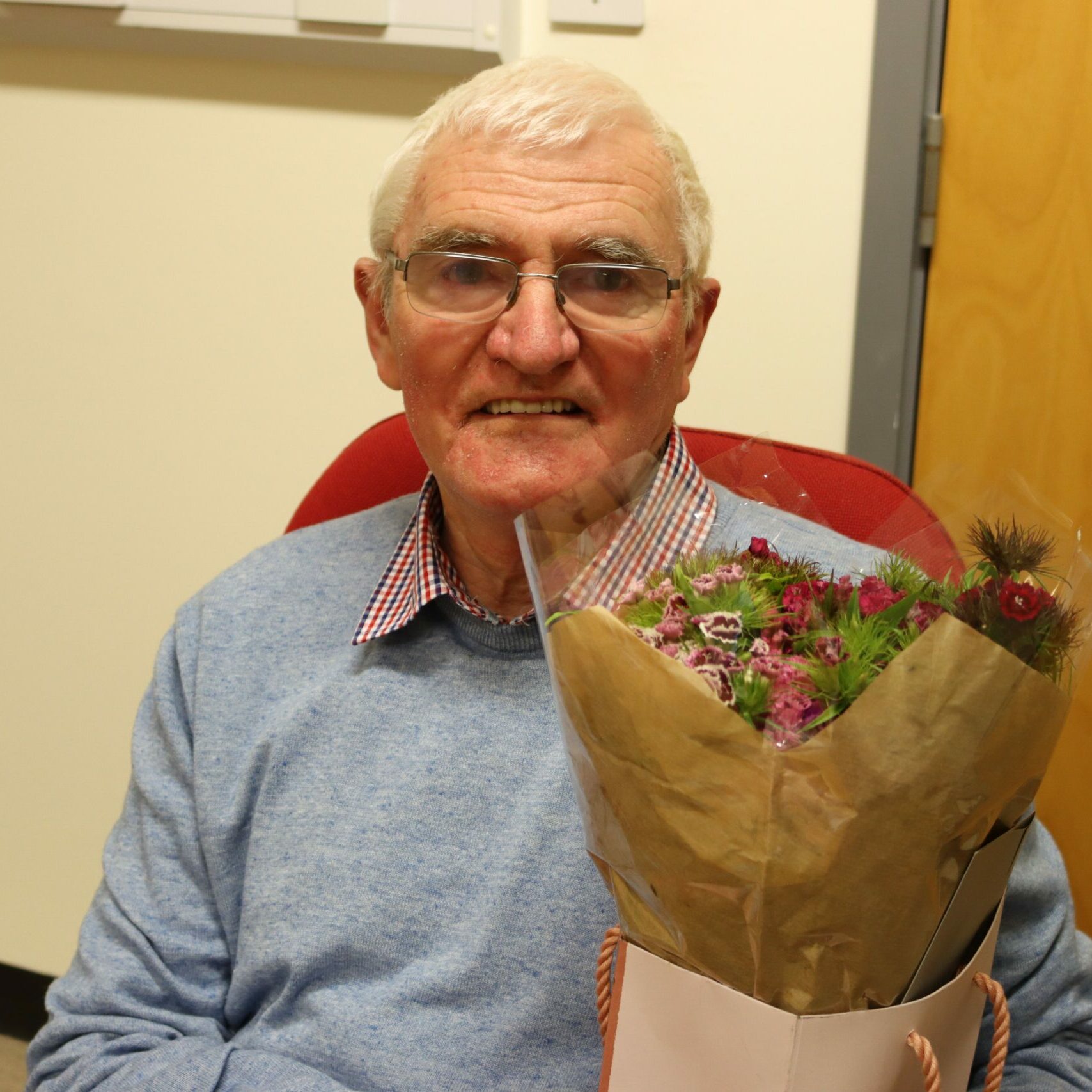 Joe Sim is sat smiling directly at the camera with a bouquet of flowers