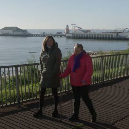 Two women walking along the seafront with a pier in the background