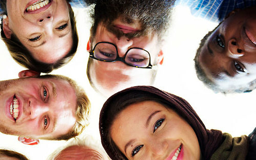 A multi-cultural group of people in a circle looking down together.