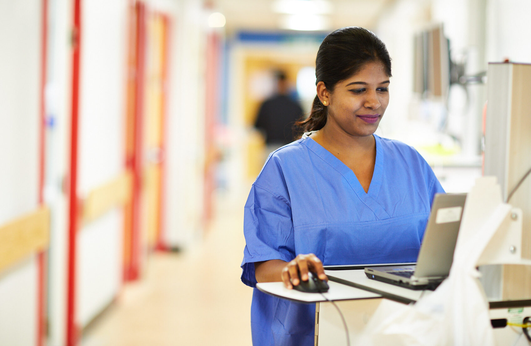 A clinician standing on a hospital ward and wearing blue scrubs works on a computer.