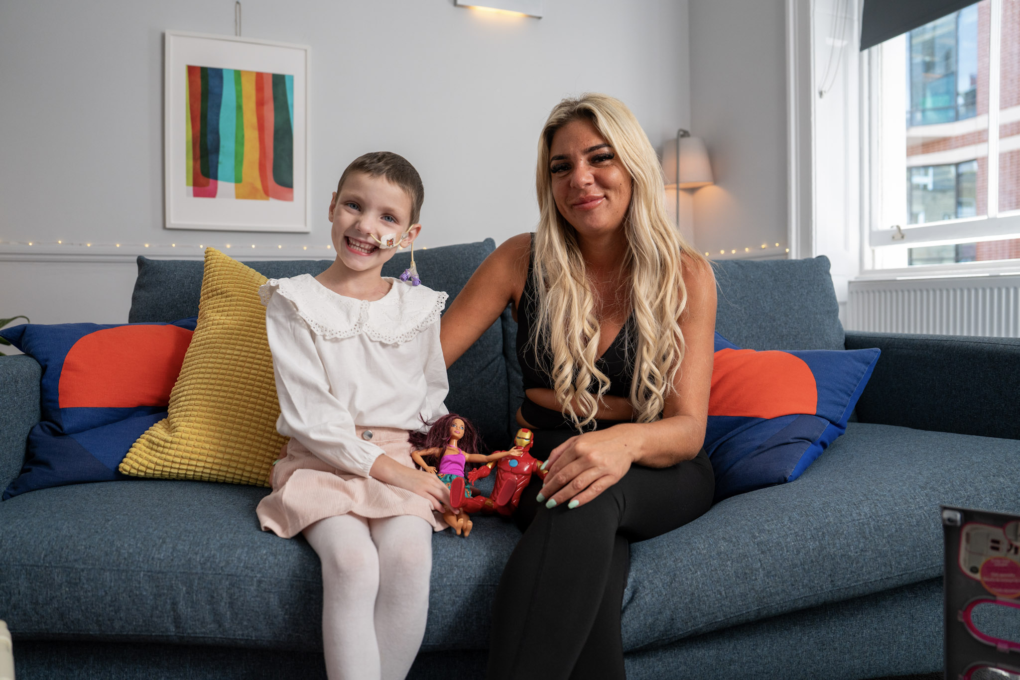 An adult and a child sit together on a sofa in a brightly lit room. The child holds two dolls while the adult rests an arm around them, creating a warm and supportive atmosphere